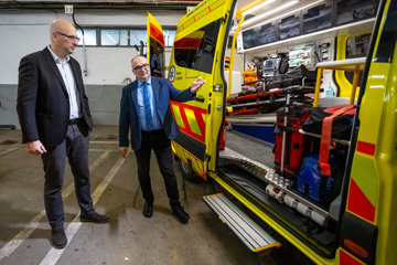 Two men in formal attire stand beside an open ambulance, showcasing medical equipment inside