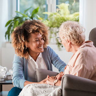 A young woman with curly hair smiles and holds hands with an elderly woman with white hair, both sitting in a bright room with plants.